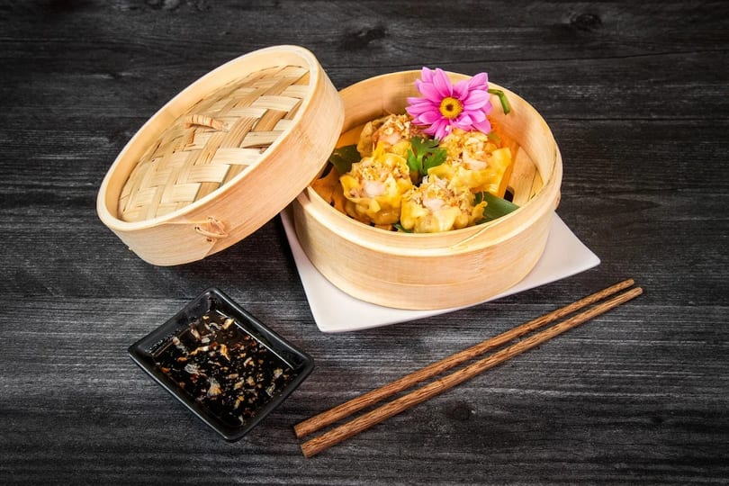 Steamed dumplings with a pink flower in a bamboo steamer, with chopsticks and sauce.