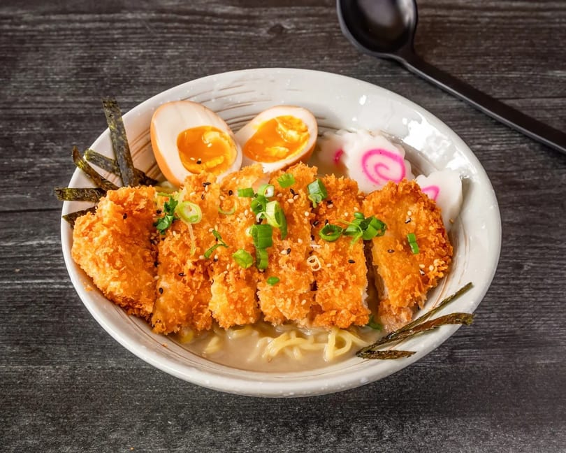 Crispy chicken katsu ramen with eggs, fish cakes, and seaweed in a white bowl.