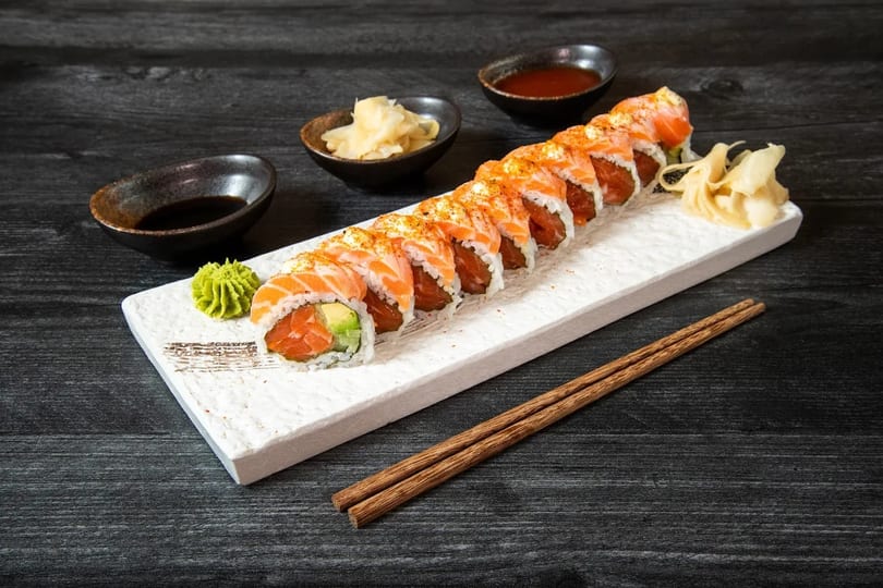 Salmon and avocado sushi roll with wasabi, ginger, and soy sauce on a white plate.