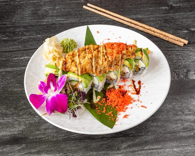 Sushi roll topped with fried tempura, avocado, and orange roe, garnished with a purple orchid.