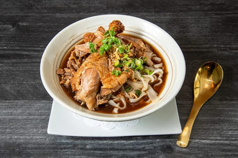 A bowl of duck noodle soup with flat rice noodles, green onions, and crispy garlic.