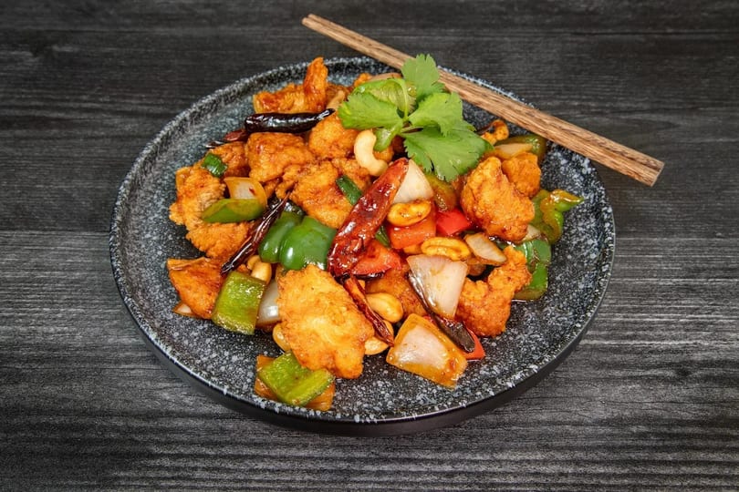 Kung Pao chicken with cashews, peppers, onions, and dried chilies on a dark plate with chopsticks.