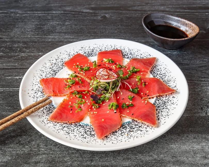 Tuna tataki with green onions, sesame seeds, and microgreens, served with chopsticks and soy sauce.