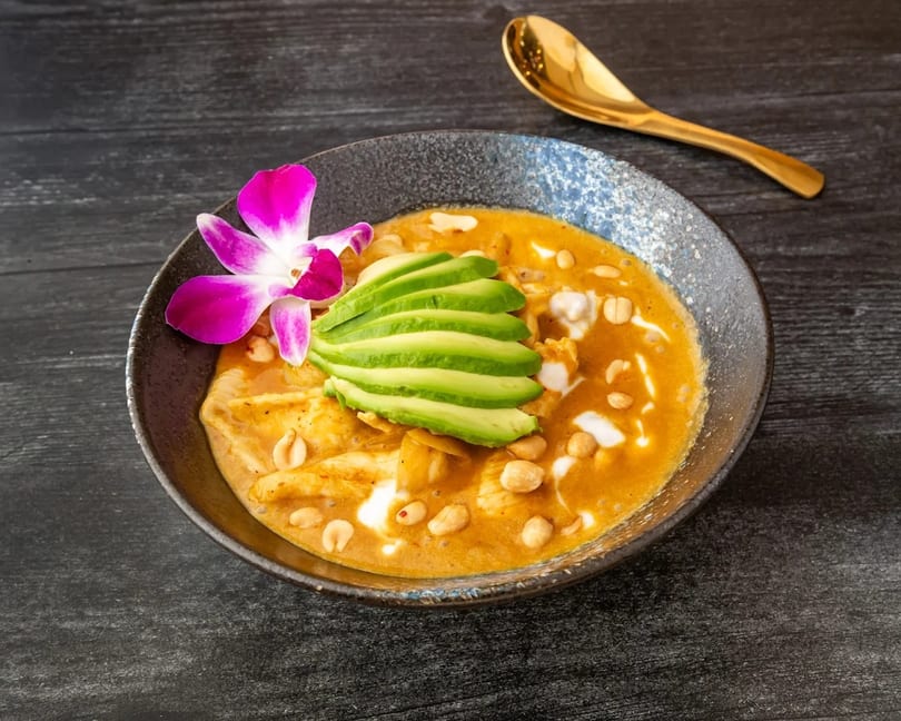 A bowl of orange curry with chicken, peanuts, avocado slices, and a purple orchid, with a gold spoon.