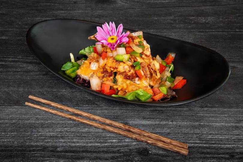 Crispy fried soft-shell crab with bell peppers, onions, and a pink flower garnish on a black plate.