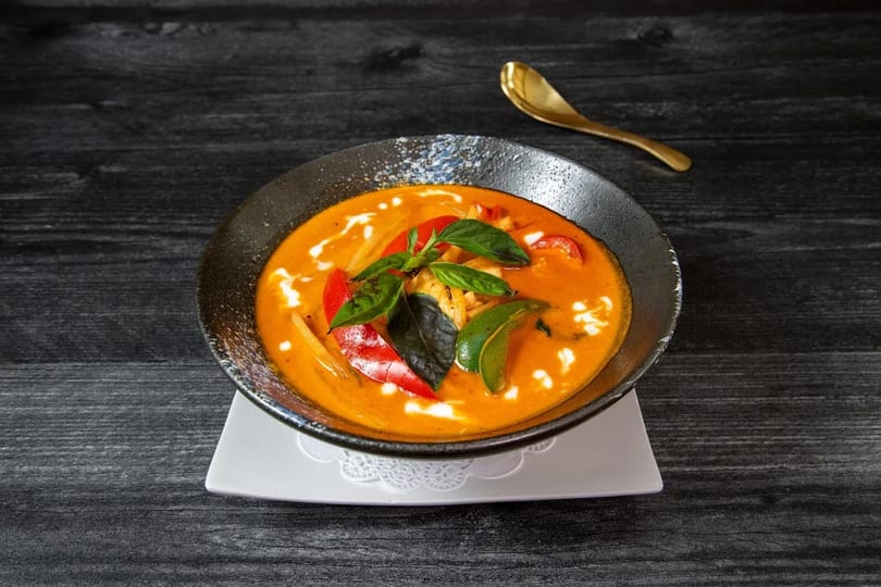Red curry with chicken, red peppers, and basil in a dark bowl on a black wooden table.
