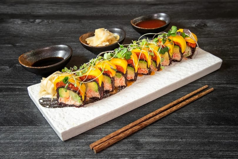 Sushi roll with mango, avocado, and crab, garnished with microgreens, on a white plate with chopsticks.