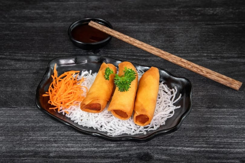 Three golden spring rolls with parsley, shredded carrots, and white noodles on a black plate.
