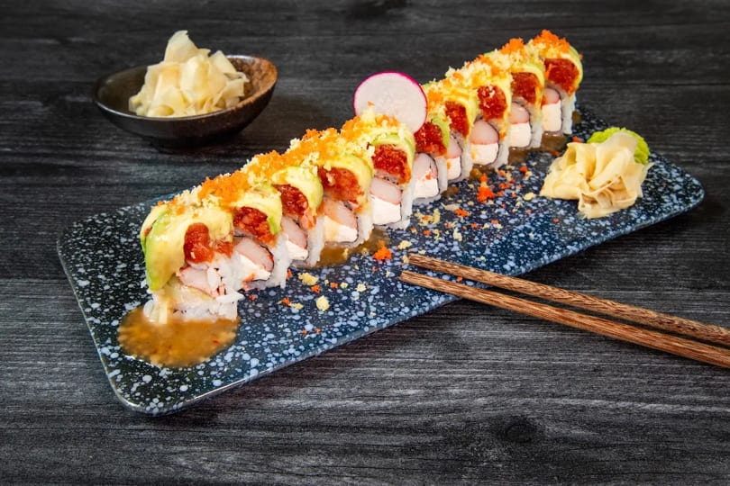 Sushi roll with avocado, red topping, and orange roe, served with ginger and chopsticks.