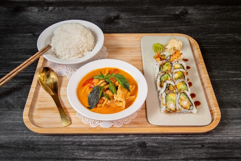 Red curry with chicken and basil, white rice, and shrimp tempura sushi rolls on a wooden tray.